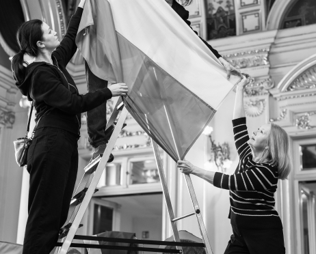 Czarno-białe zdjęcie. Natalia Strzelecka stoi na drabinie i obiema rękami unosi dużą flagę, przygotowując ją do zawieszenia. Bożena Adamek i Ania Syrbu pomagają jej, podtrzymując materiał z boku i od dołu. W tle widoczne są dekoracyjne sklepienia wnętrza teatru oraz ustawione krzesła.